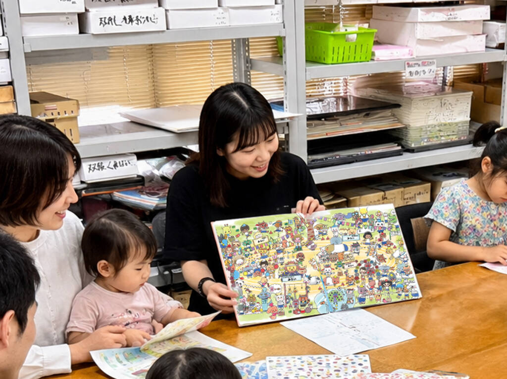 お母さんに抱かれた小さな子どもが、開かれた絵本に手を伸ばしている様子