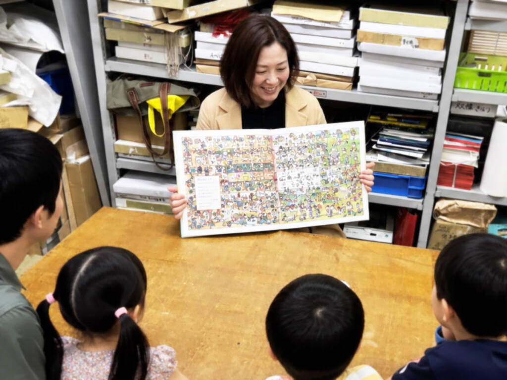 飯塚印刷の社屋で、紙の見本が並んだ棚を背景に、絵本の読み聞かせに聞き入る子どもたち